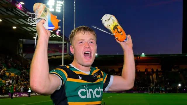 Northampton Saints fly-half Fin Smith holding his boots up in celebration after the final whistle.