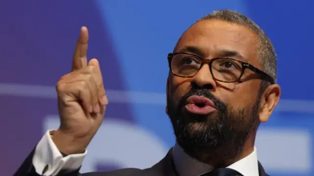 Shadow housing secretary James Cleverly wears black-rimmed glasses and a suit, he gestures with one finger pointed in the air while speaking