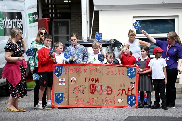 Schoolchildren hold massive hand-made cardboard banner which says "Thank you Jill from St Hilds" and "Go Jill go". There are flanked by teachers on either side.