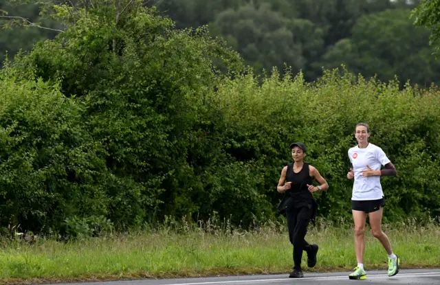 Jill Scott running on a path with hedges behind her. She is wearing a white top and green trainers.
