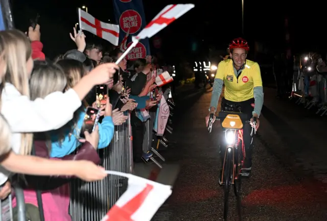 Jill Scott wearing yellow top and red helmet cycling in the dark. People with England flags line up on barriers on the left hand side cheering her on.
