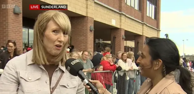 Jilll Scott's mum is interviewed by BBC presenter Nisha Joshi outside the Stadium of Light. She has shoulder length blonde hair and is wearing a light brown jacket. Nisha is holding a black microphone with a white BBC logo on it