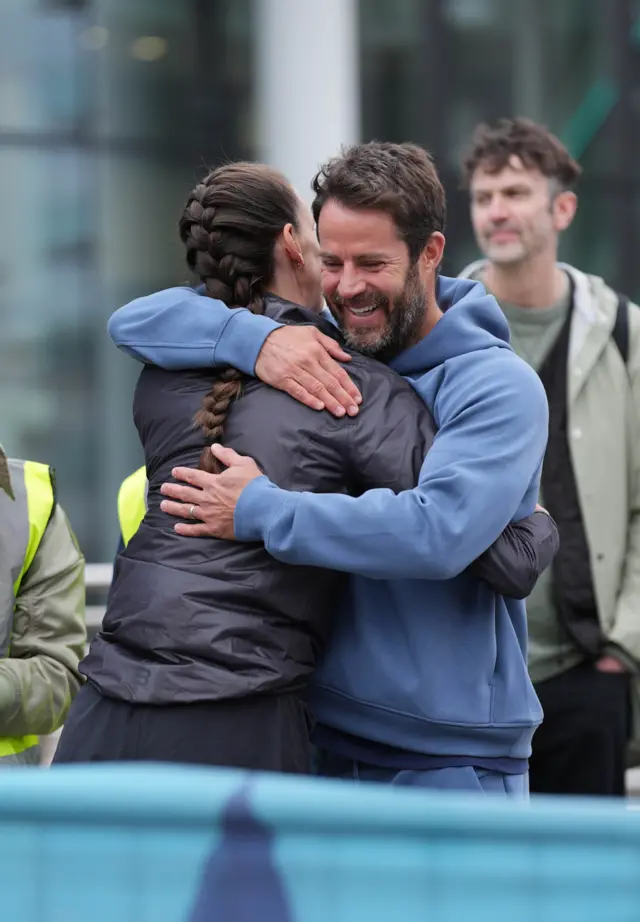 Jill Scott hugs Jamie Redknapp. She is wearing black clothes and her hair is in a plait down her back. Jamie Redknapp is wearing a light blue tracksuit