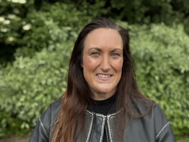 Close up photo of Amanda Wilson who has long brown hair and is wearing a black jacket. She also has a nose ring and is smiling. There are green hedges in the background.