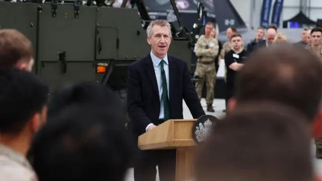 Dan Jarvis stands at a lecturn at a military facility surrounded by men in camouflage fatigues