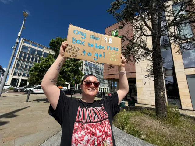 Chloe Rose is wearing a black Lioness top with red lettering and is wearing sunglasses. She is holding a cardboard sign above her head which says "Only about 90 box to box runs to go!"