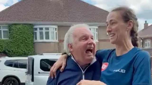 Adam Walter and Jim Scott in front of a house. Scott has her arm around Walter who is looking at her smiling with an open mouth.
