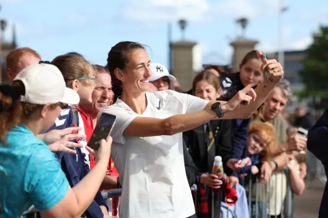 Jill Scott takes a selfie with supporters outside the Stadium of Light. She is wearing a whit t-shirt and is holding a phone in her hand and standing in front of a line of people