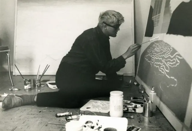 A black and white photo of Hockney painting kneelig on the ground his home studio in Los Angeles in 1966