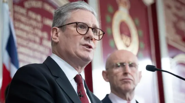 Starmer in front of a microphone, with Healey looking on behind him