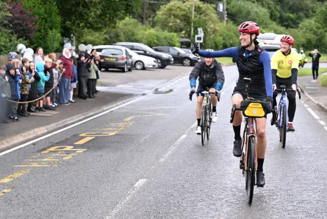 Jill Scott in blue cycling gear and red helmet smiling and looking at crowd on pavement. She is on a bicycle on the road flanked by two cyclists behind her.