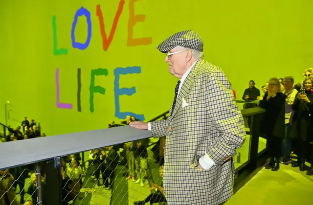 David Hockney, wearing a chequered suit and flat cap, is seen looking over a balcony at the gala opening of an exhibition