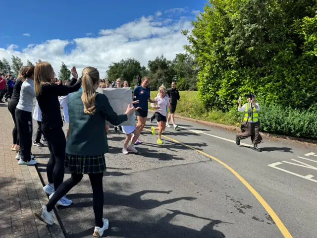 Sunny day with lots of pupils in school uniform holding signs as Jill Scott runs on road