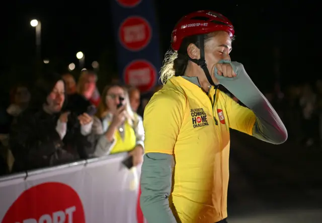 Jill Scott looks emotional as she puts her hand to her mouth. She is wearing a yellow cycling top and a red helmet. There are people in the crowd behind her taking photos.