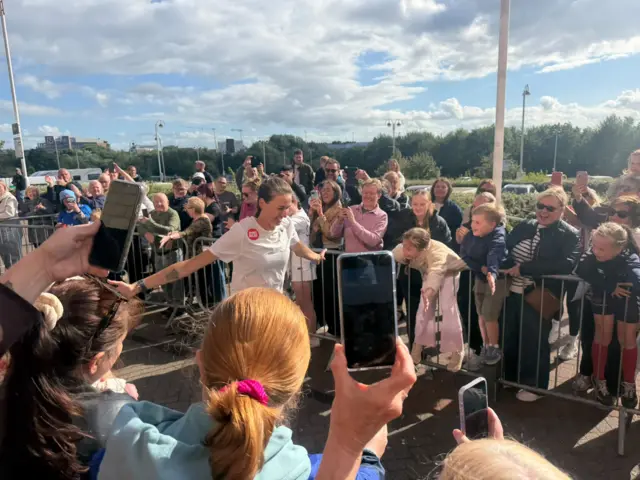 Jill Scott smiling as she runs through crowd with many of them holding their phones as they take photos of her. It is a sunny day.