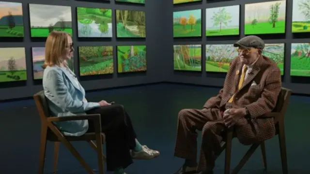 Katie Razzall wearing a blue blazer, speaking to David Hockney, wearing a cap and checked blazer, They are both sitting down.