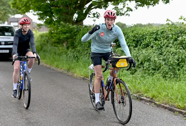 Jill Scott in light blue cycling gear on a cycle. She is looking at camera in a victorious way with a fist bump. She is flanked by a cyclist behind her in grey cycling gear.