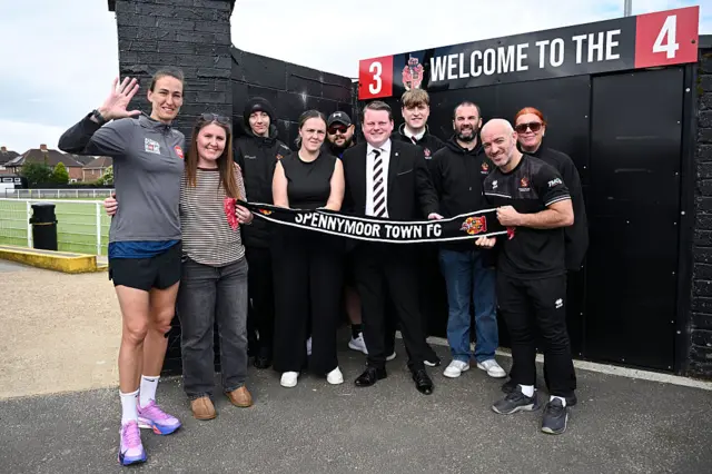 Jill Scott posing with nine people in front of a football stadium. There are all holding one scarf which says "Spennymoor Town FC". They are all smiling and posing to the camera.