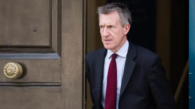 Dan Jarvis, wearing a dark suit, white shirt and maroon tie, walks out of a brown doorway