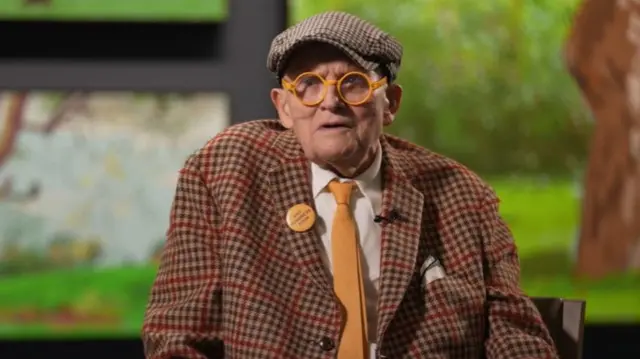 David Hockney, wearing a flat cap, orange glasses, tie and checked blazer.