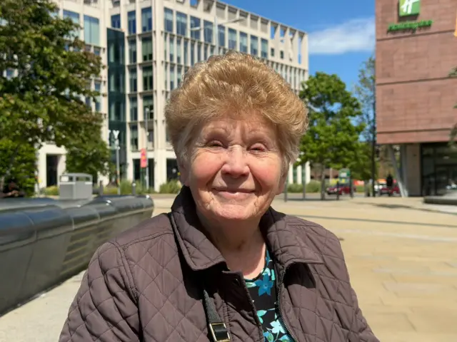 Kathleen Boddy sitting in Kele Square in Sunderland. She has short light brown hair and is smiling. She is wearing a brown padded jacket and a blue and black floral shirt underneath