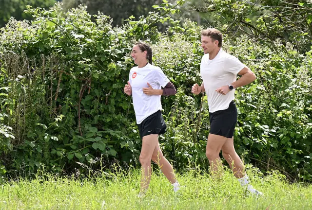 Jill Scott running with ultrarunner Jonny Davies. They are both wearing black shorts with white T-shirts. Jill Scott's has the Sport Relief logo on it which is a red circle with the words in white letters. There is long grass in the foreground and bushes in the background