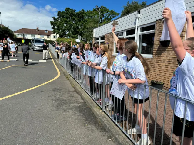 Sunny day with lots of pupils in school uniform holding signs behind railing and waving
