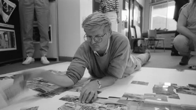 Black and white photo of Hockney making a photo collage on the floor.