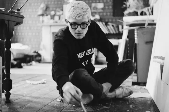 Hockney, pictured at work in his studio in the 1960s with a paint brush in his hand, in a black and white image