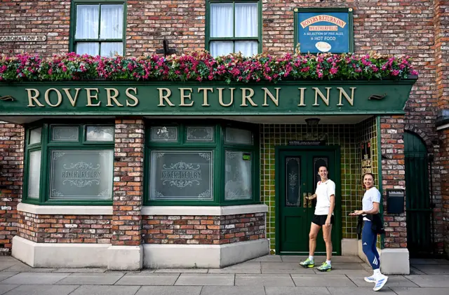 Jill Scott and Dame Jessica Ennis-Hill on the Coronation Street set. Jill is wearing black shorts, a white T-shirt and has one hand on a door handle. Jess is wearing a white top, blue leggings and white trainers and is half turned to the camera. The set is a red brick with the word Rovers Return Inn in gold on a green background