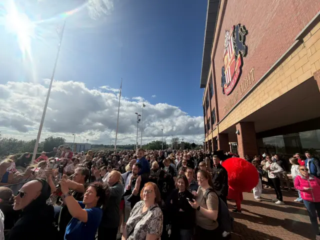Hundreds outside stadium in Sunderland shirt on a sunny day.