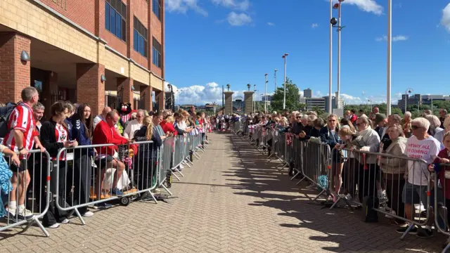 Crowds standing behind barriers on a sunny day by the finish line.