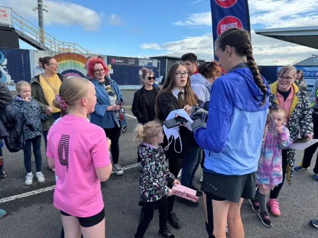 Jill Scott in blue jacket signs autograph. She is surrounded by pupils and adults. It is a sunny day.