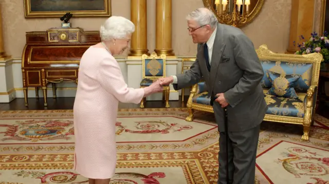 David Hockney, wearing a grey suit, shaking the hand of Queen Elizabeth II, who is wearing a pink outfit.