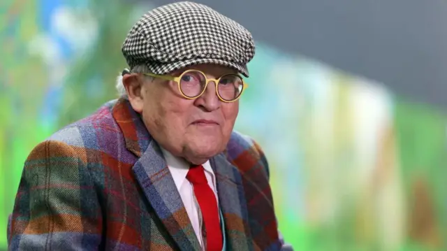 David Hockney pictured in a check jacket in front of one of his paintings