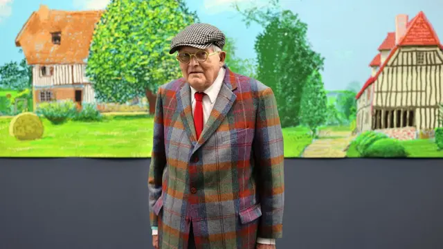 David Hockney, wearing a checked pattern blazer and red tie, is standing in front of a colourful landscape painting of Normandy.