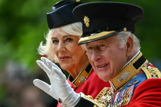 The King waves as he arrives at the Trooping the Colour. In his background Queen Camilla can be seen next to him also smiling. Both of them are in full military attire