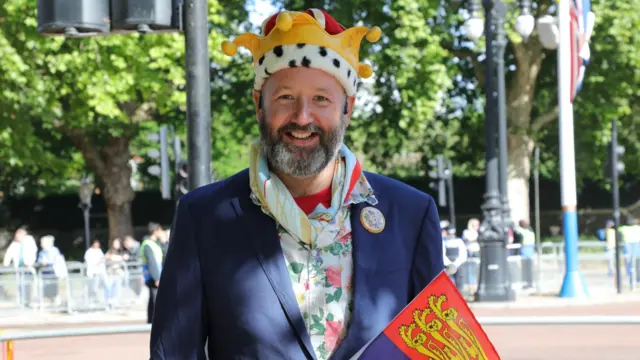Corrigan Gommenginger stands in a suit and holds the royal standard flag. He is wearing a plush novelty crown.