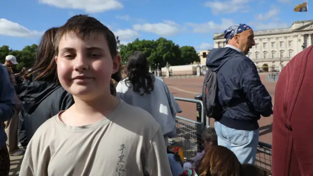 Owen stands behind a barricade at the palace in a beige T-shirt. he is a boy of about 12.