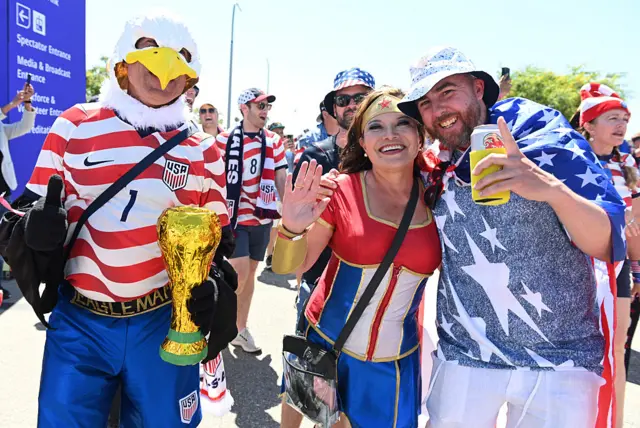 A man wearing an Eagles head and holding a World Cup stands next to a woman wearing an American branded outfit