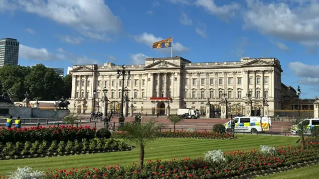 The Royal Standard flag can be seen flying above Buckingham Palace, with the lawn in the foreground. Police vehicles and security are also in view.