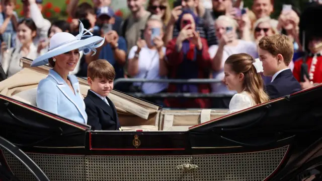 The Princess of Wales and her three children sit in a carriage during the Trooping the Colour. Catherine is seen wearing a pale blue jacket and hat