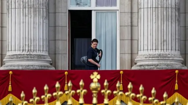 A palace staff member cleans the balcony. she is holding a hoover and can be seen working behind the red drapes hung over the balcony.
