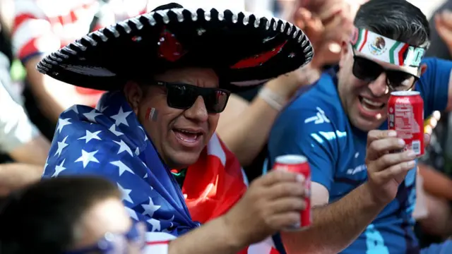 A fan wearing a wide brimmed hat with a usa flag round his shoulders celebrates with a coke in his hand.