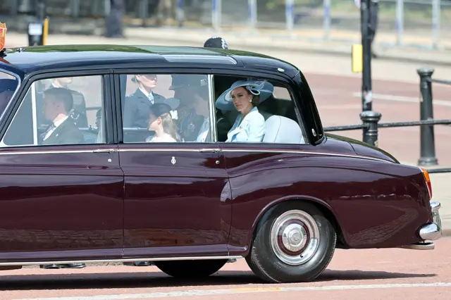 The Prince and Princess of Wales can be seen in a car arriving at Buckingham Palace. The back window is wound down and Princess Catherine is looking out, wearing a blue hat and jacket.
