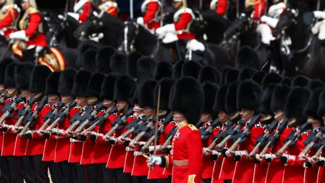 Troops stand on display in their red uniforms and bearskin hats as they take part in the Trooping the Colour. They are seen holding their rifles