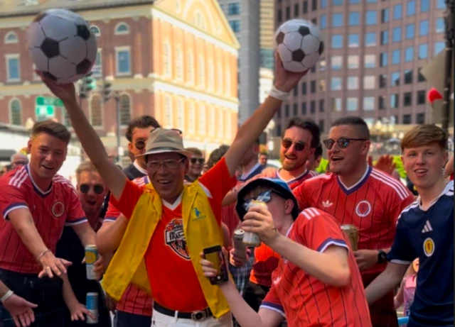 a man holding two footballs poses for a picture with several Scotland fans