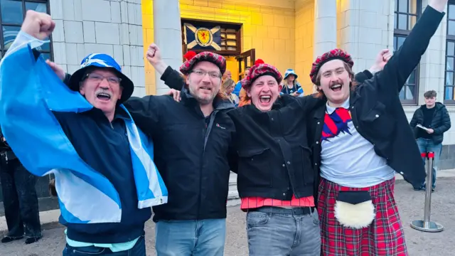 Four Scotland fans celebrating the win outside Aberdeen's Beach Ballroom, one is wearing a kilt, another is draped in a Saltire flag.