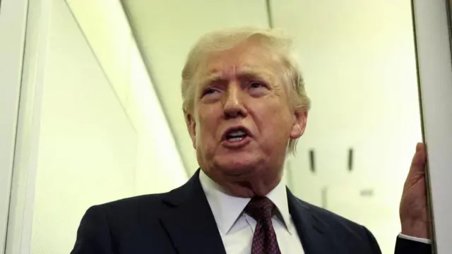 US President Donald Trump wears a suit and holds onto a wall as he speaks to to reporters aboard Air Force One in March
