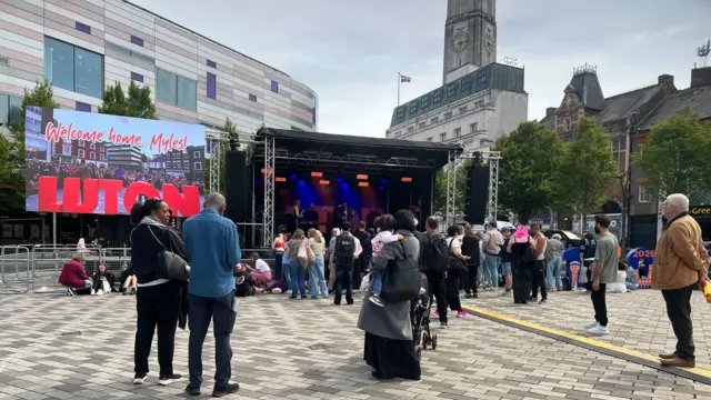 People are gathered in a large square in front of a stage. A screen says "welcome home Myles" on it next to the stage. Overlooking the square is a large clock tower.